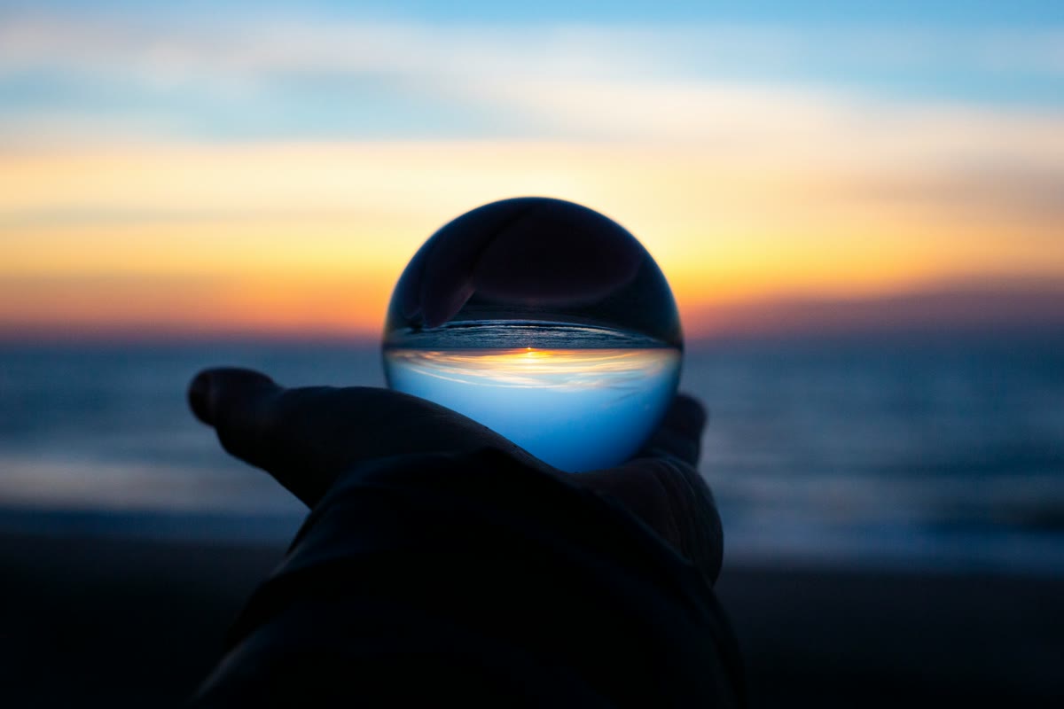 Hand holding a crystal ball reflecting an inverted sunset over the ocean